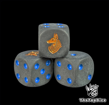 Close up of a matte black 16mm resin D6 die engraved with a gold anubis jackal and blue pips