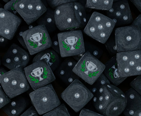 Pile of matte black second place trophy dice with silver pips and silver number two cups in a green laurel